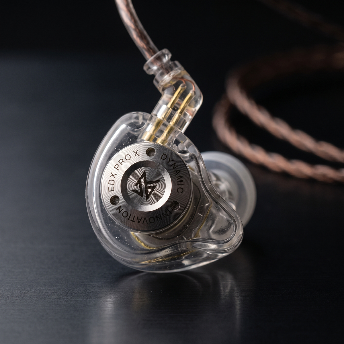 A close-up of a transparent in-ear monitor earpiece with a silver metal center ring engraved with 'EDX PRO X' and 'DYNAMIC INNOVATION', attached to a braided copper cable on a dark reflective surface.